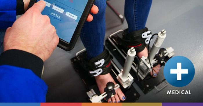 A person’s hands are shown holding a tablet with an application open and a patient’s legs strapped into a complex mechanical device